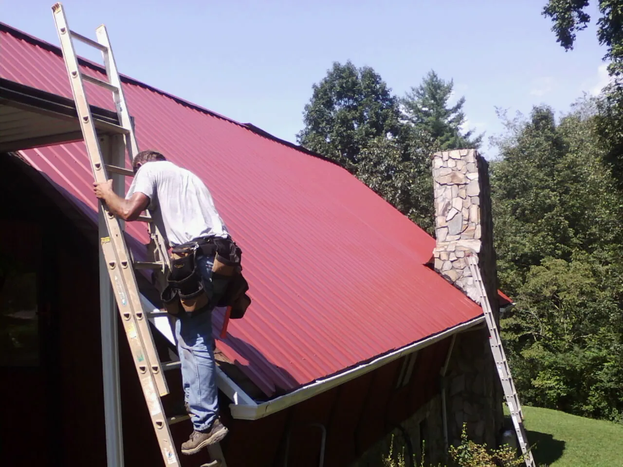 Roofer on a ladder beside a red metal roof and stone chimney — Soffit & Fascia Repair by Roofing Brothers in Centennial
